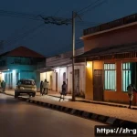 기니비사우에서 안전하게 여행하는 법 - A detailed urban street scene in Bissau, Guinea-Bissau at dusk, showing a cautious traveler walking ...
